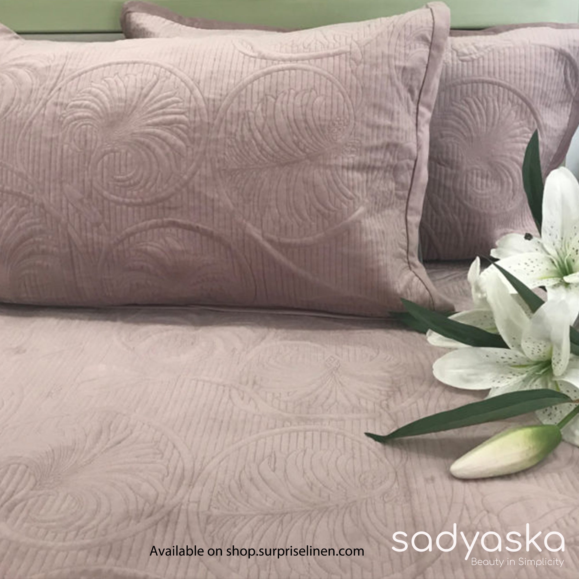 A onion pink velvet bed cover set with a floral pattern, displayed on a bed with a white lily flower on the corner, and a text indicating the brand Sadyaska was present.